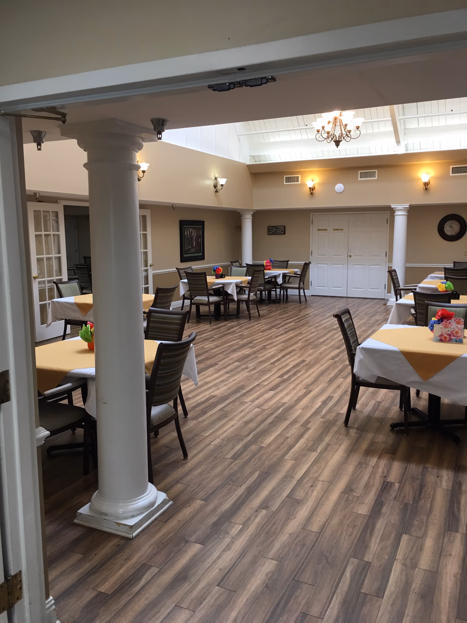 Interior view of a dining room with several tables covered with white and yellow tablecloths, each decorated with colorful flower centerpieces. The room features wooden flooring, beige walls, white columns, wall sconces, a chandelier, and a clock on the wall.