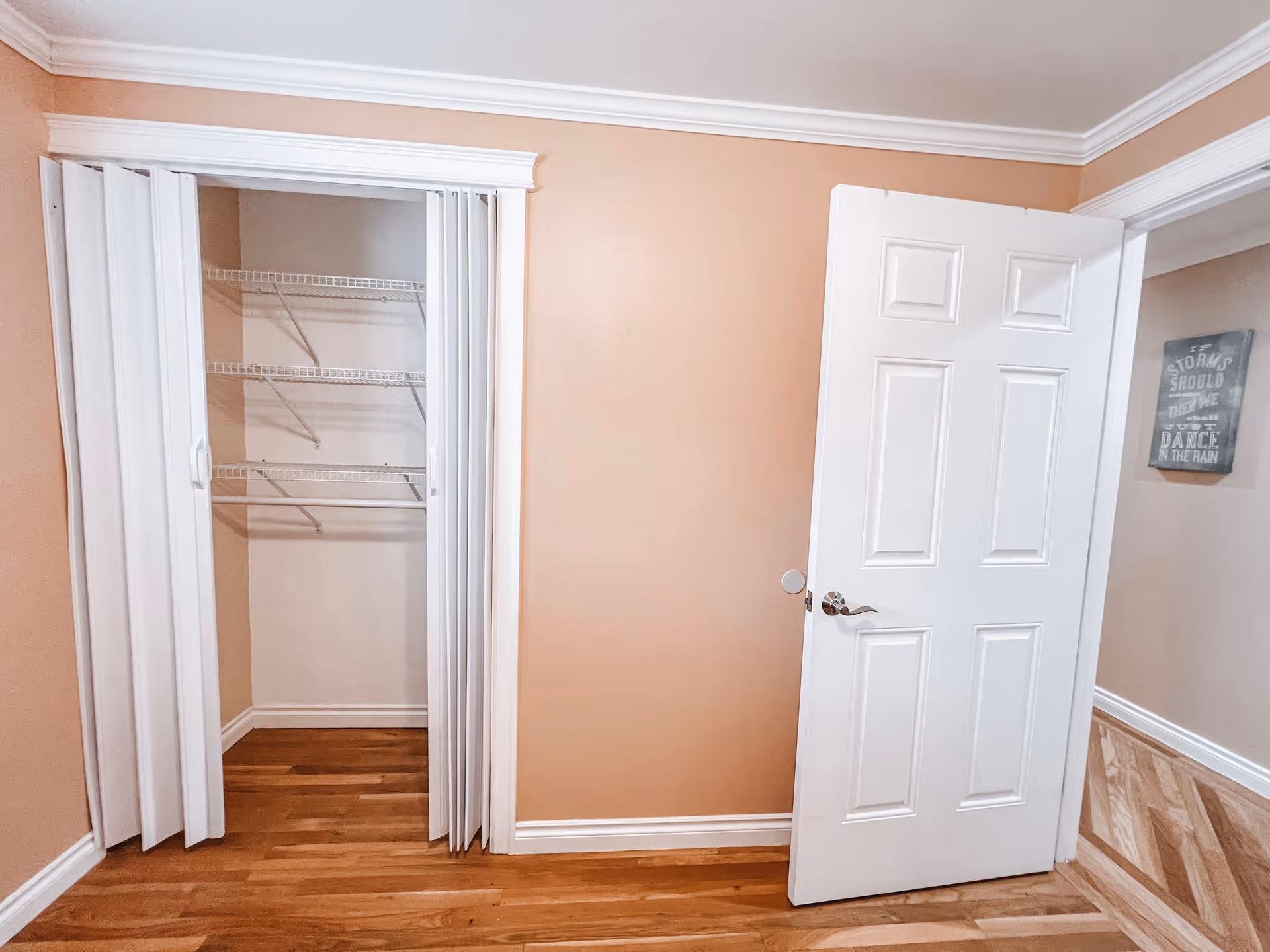 An empty room with light brown walls and wooden flooring. There is a white folding closet door partially open revealing wire shelving inside. A white door is open leading to another room with a framed sign on the wall.