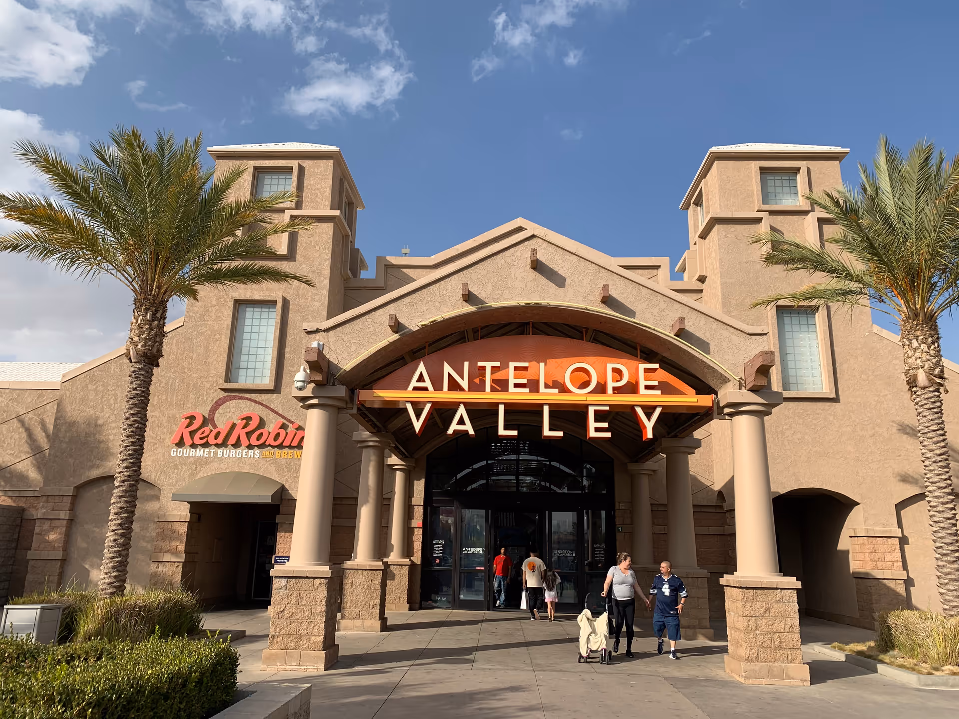 Entrance to a shopping center named Antelope Valley with a Red Robin restaurant visible on the left side. The building has a beige stucco exterior with two palm trees flanking the entrance. Several people are walking near the entrance under a clear blue sky.