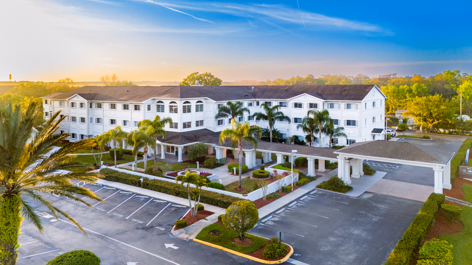 Exterior view of a large, white three-story senior living facility building with a covered entrance and surrounding palm trees and landscaping under a clear blue sky at sunset.