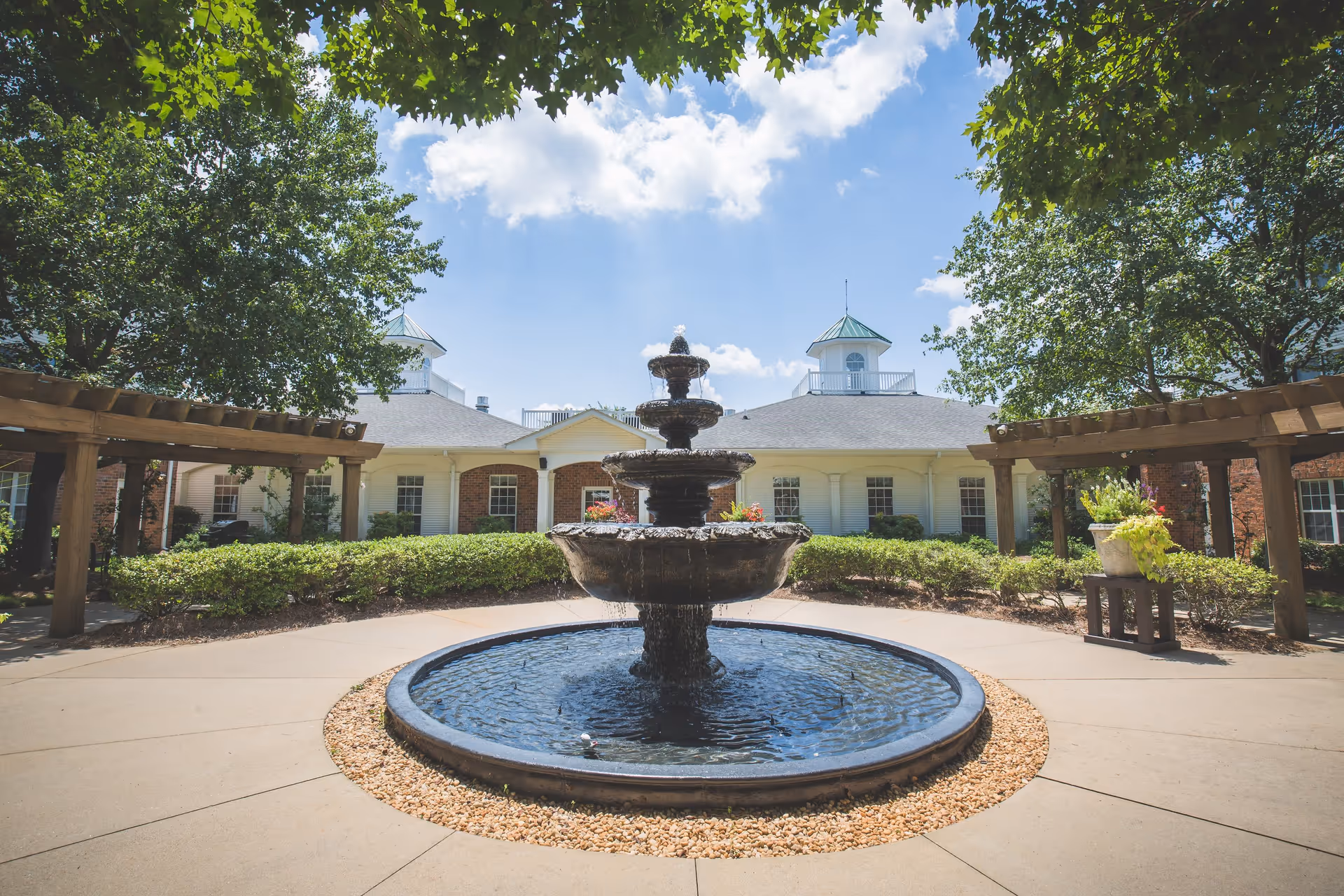 A three-tier stone fountain in a landscaped courtyard in front of a brick-and-white senior living building under a blue sky.
