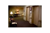 Interior hallway of a senior living facility with carpeted floor, beige walls, and several closed doors. There are chairs along the left wall and a table with a lamp and framed pictures at the end of the hallway.