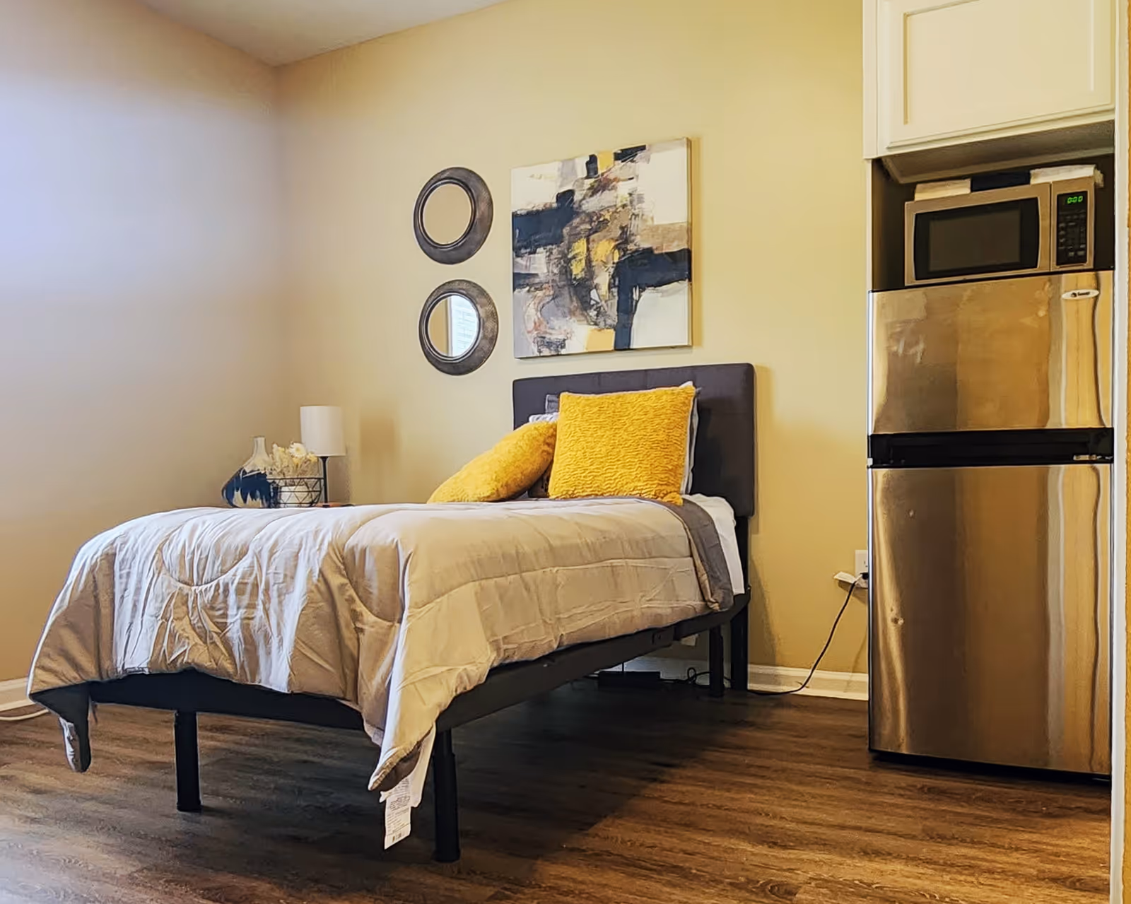 A small bedroom with a single bed featuring a gray headboard, beige comforter, and two yellow pillows. On the wall above the bed are two round mirrors and an abstract painting. To the right of the bed is a stainless steel mini refrigerator with a microwave on top. The room has wooden flooring and light-colored walls.