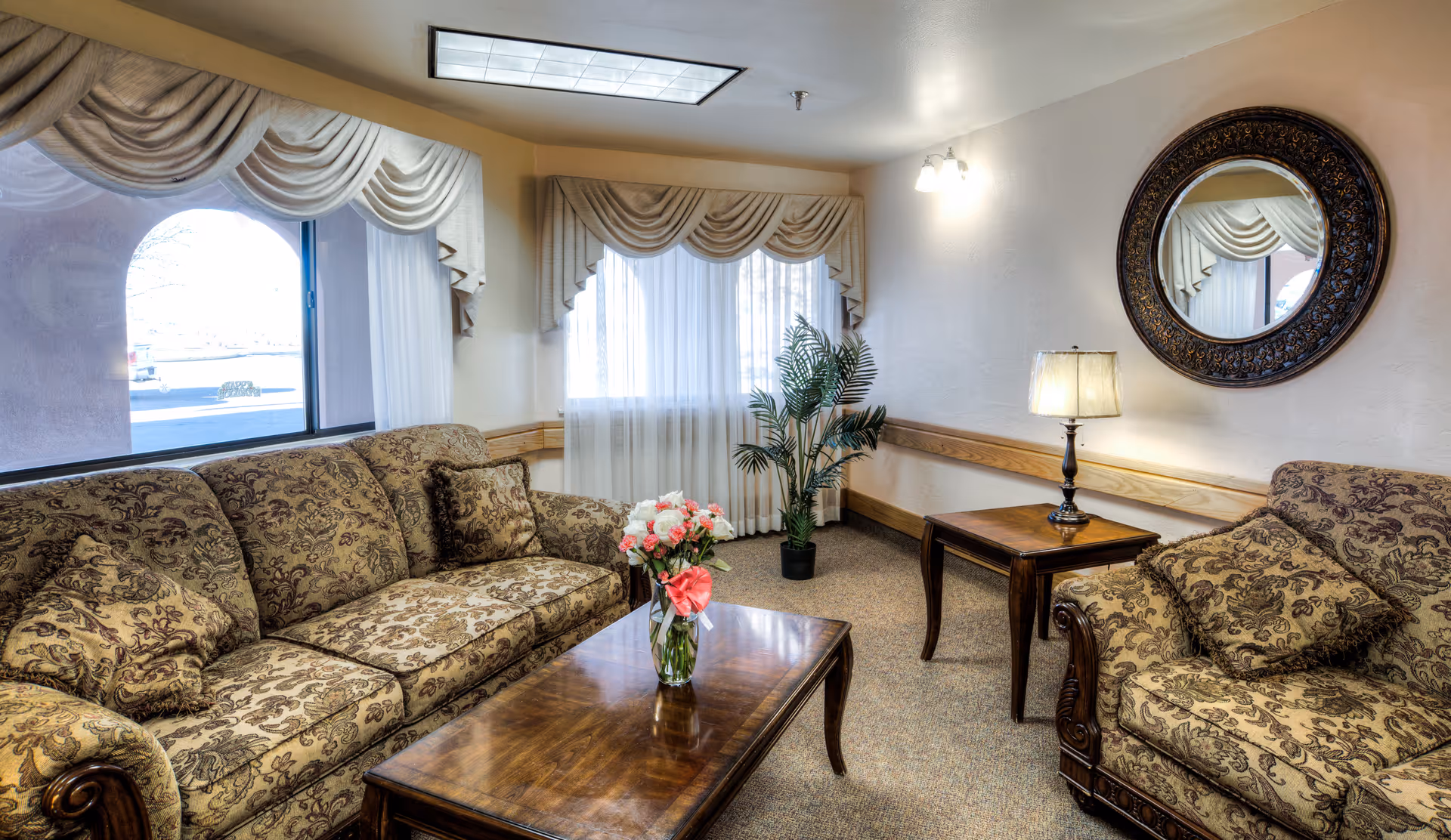A cozy living room area with two patterned sofas, a wooden coffee table with a vase of flowers, a side table with a lamp, a large round decorative mirror on the wall, and windows with sheer curtains and valances. There is also a potted plant in the corner.