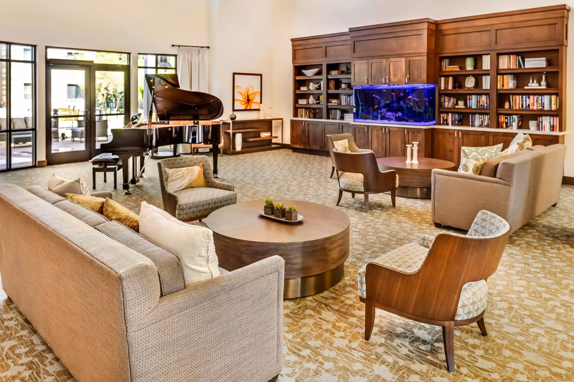 A spacious and well-lit living room area featuring two beige sofas, several cushioned armchairs, and two round wooden coffee tables. A grand piano is positioned near large glass doors that lead outside. The back wall has built-in wooden shelves filled with books and decorative items, and a large aquarium with blue lighting is integrated into the shelving unit. The room has a patterned carpet and light-colored walls.