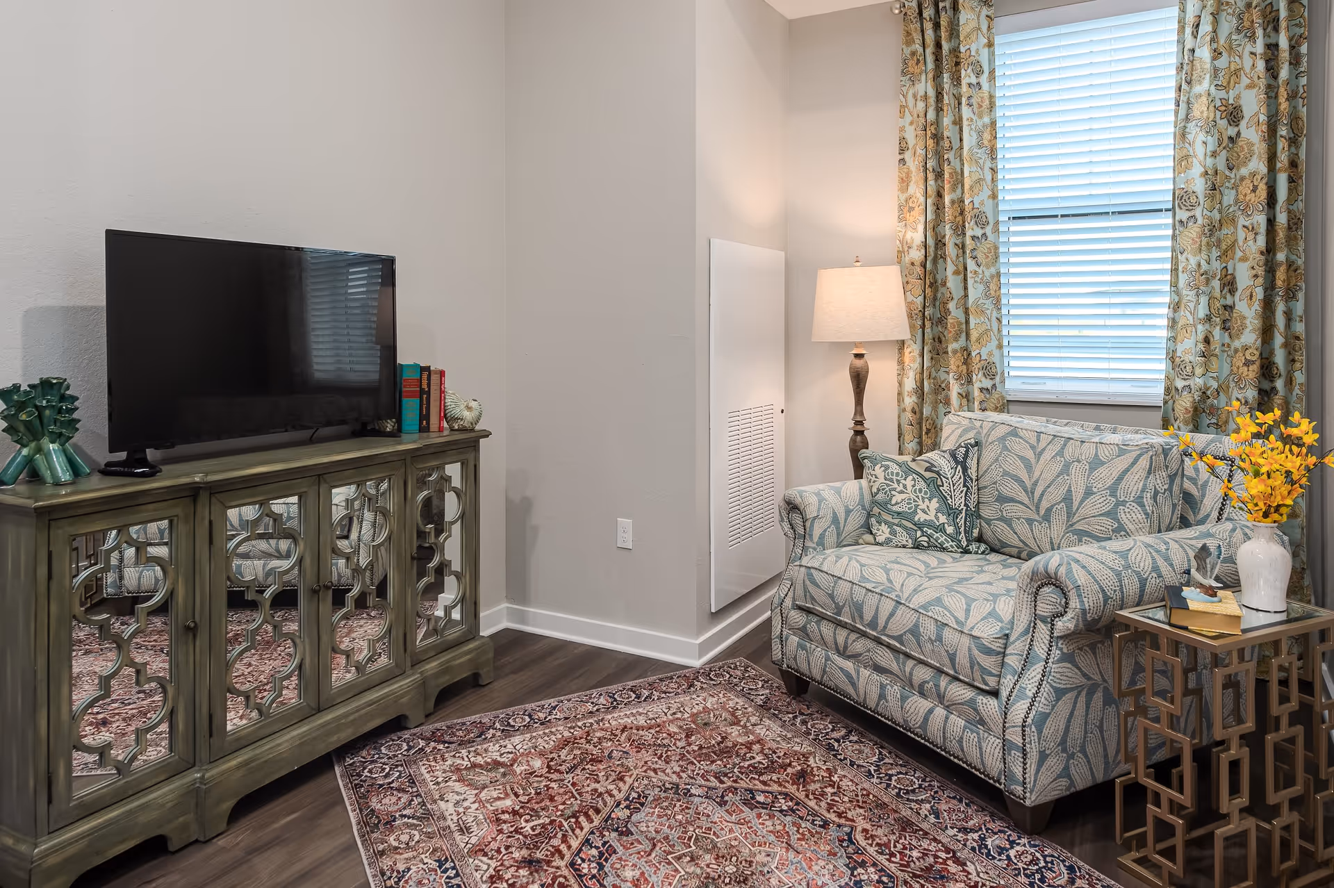 A cozy living room corner featuring a patterned loveseat with matching cushions, a floor lamp, and a window with floral curtains. Next to the loveseat is a decorative side table with a vase of yellow flowers and books. Across from the loveseat is a wooden cabinet with mirrored doors holding a flat-screen TV and decorative items on top. The floor is covered with a richly patterned area rug.