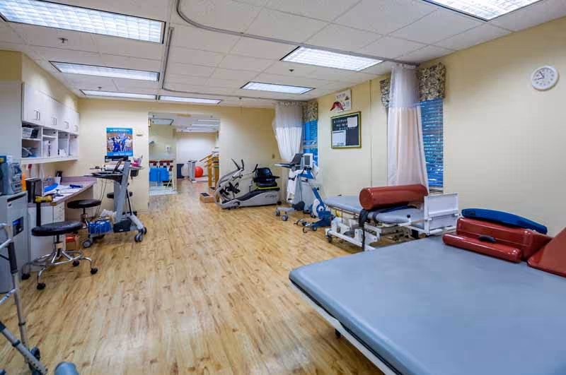 A physical therapy room with exercise equipment including stationary bikes, therapy tables with red and blue cushions, a desk with a computer, and wooden flooring. The room has beige walls, fluorescent ceiling lights, windows with curtains, and a clock on the wall.