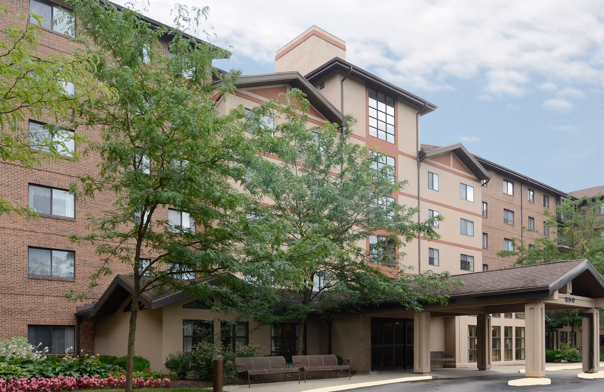 Exterior view of a multi-story senior living facility building with a covered entrance, surrounded by trees and landscaped greenery under a partly cloudy sky.