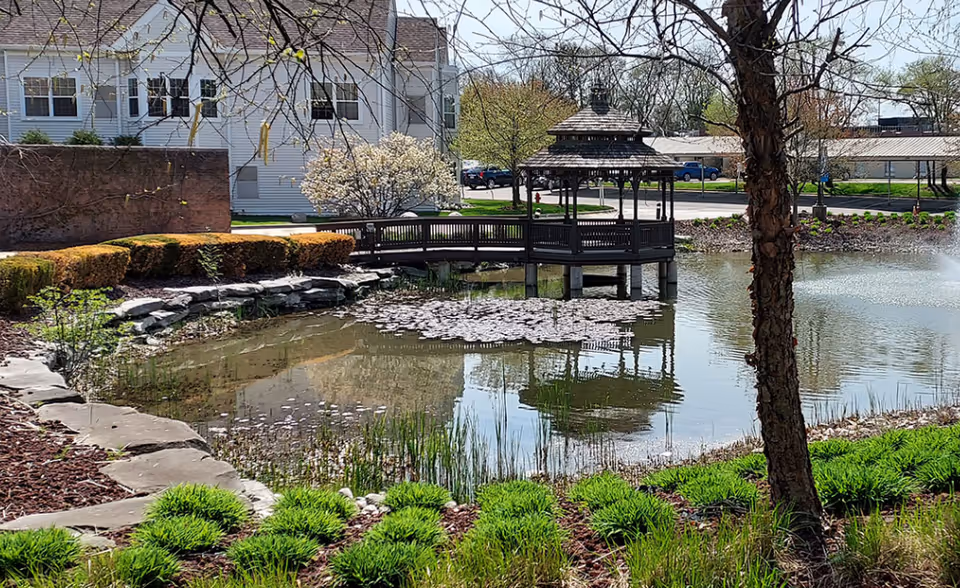 A serene outdoor scene at Autumn House Bloomfield Hills featuring a small pond with lily pads, a wooden gazebo on stilts extending over the water, surrounded by green shrubs, trees, and a stone border. In the background, there are buildings and parked cars under a clear sky.