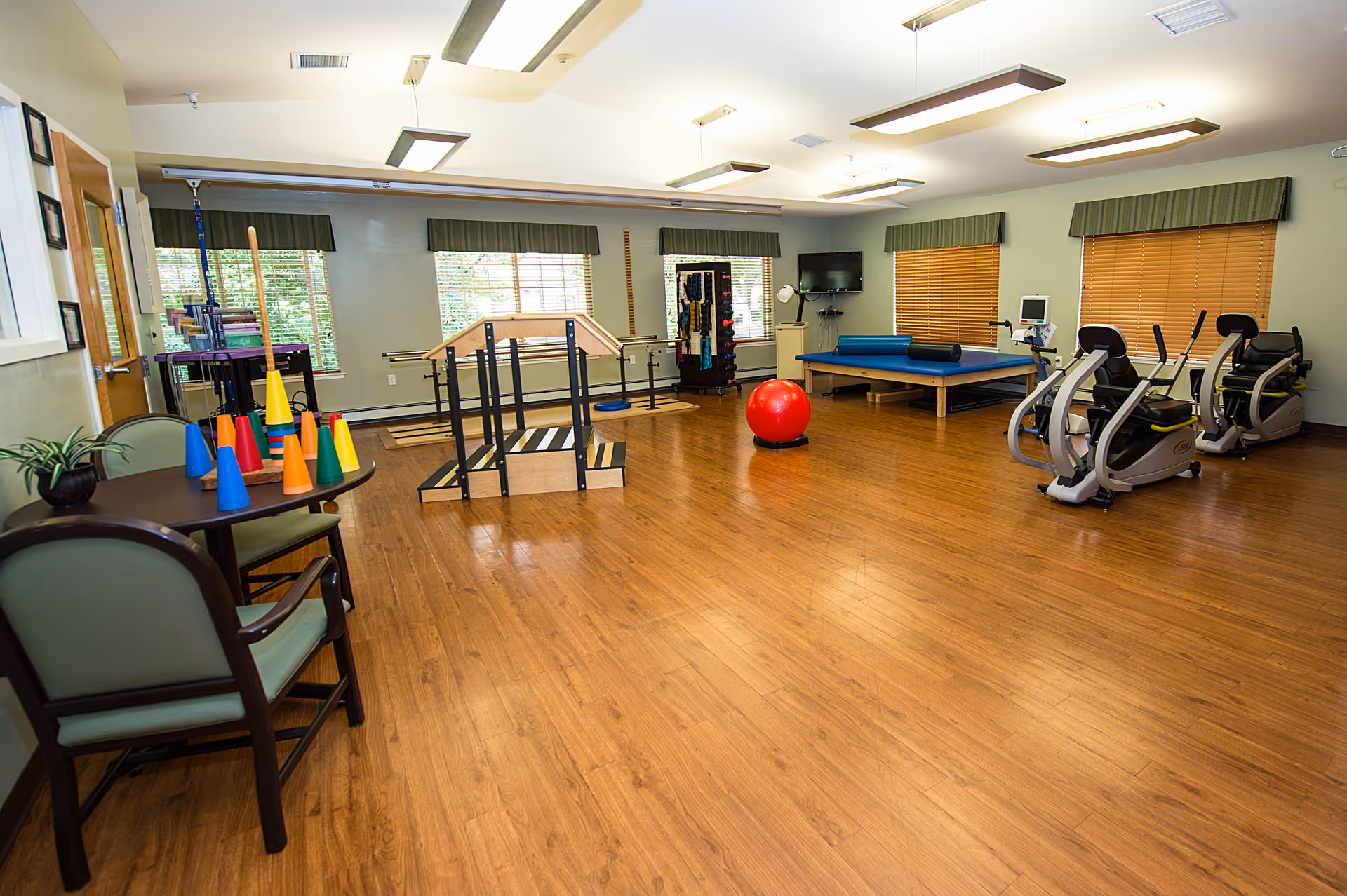 A spacious rehabilitation or physical therapy room with wooden flooring, exercise equipment including stationary bikes, a red exercise ball, parallel bars for walking practice, and a therapy table. There are windows with blinds and valances, a small table with chairs and colorful cones, and a TV mounted on the wall.