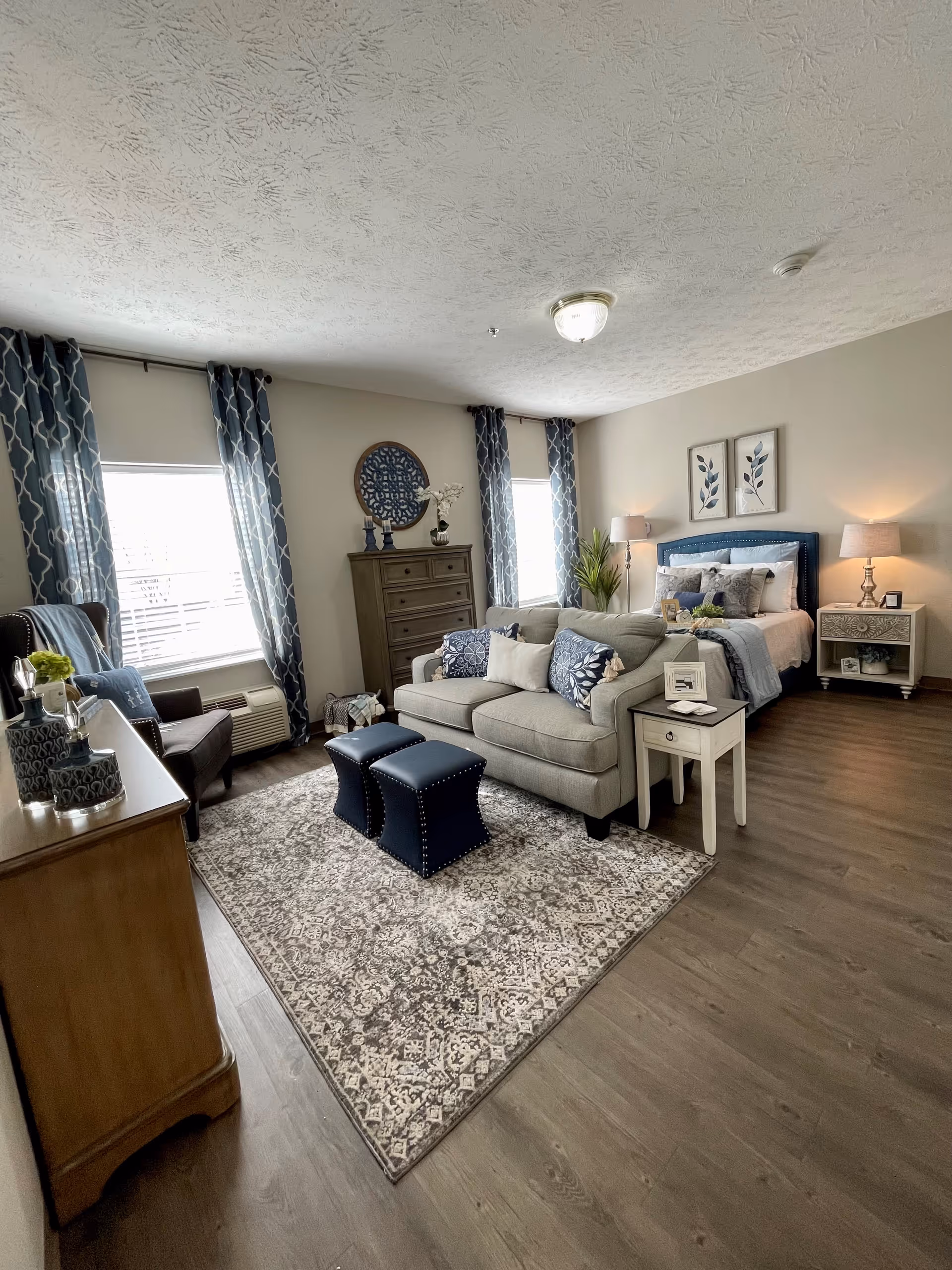 A cozy and well-decorated bedroom with a bed, a beige sofa with decorative pillows, two blue ottomans, a wooden dresser, an armchair, two windows with blue patterned curtains, a side table with a lamp, and framed botanical artwork on the wall.