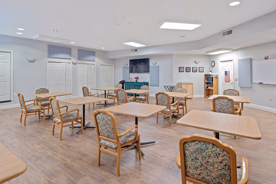 Well-lit community dining/activity room with multiple wooden tables and upholstered chairs arranged on a wood floor, a TV and storage cabinets along the far wall.