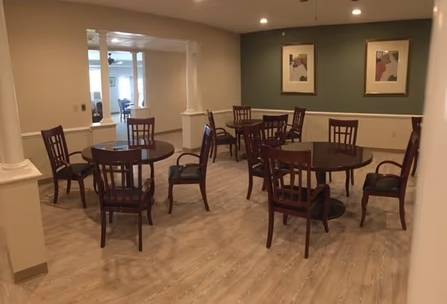 Interior view of a dining area with several round wooden tables and matching chairs arranged on a light wood floor. The walls are painted beige and green, with two framed abstract artworks hanging on the green wall. The room is well-lit with recessed ceiling lights, and there is an open doorway leading to another room in the background.
