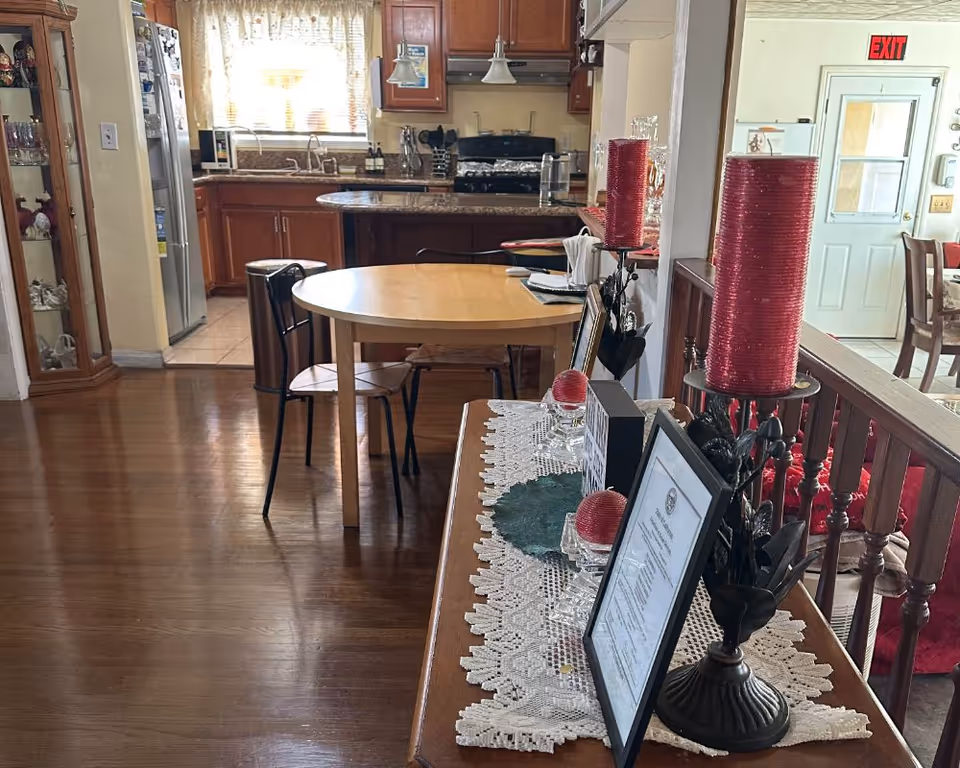 Interior view of a senior living facility showing a wooden dining table with chairs in the foreground, a kitchen area with wooden cabinets, a refrigerator, and a stove in the background. To the right, there is a decorative table with red candles, framed certificates, and other decorative items on a lace runner. An exit sign and a door are visible further back.