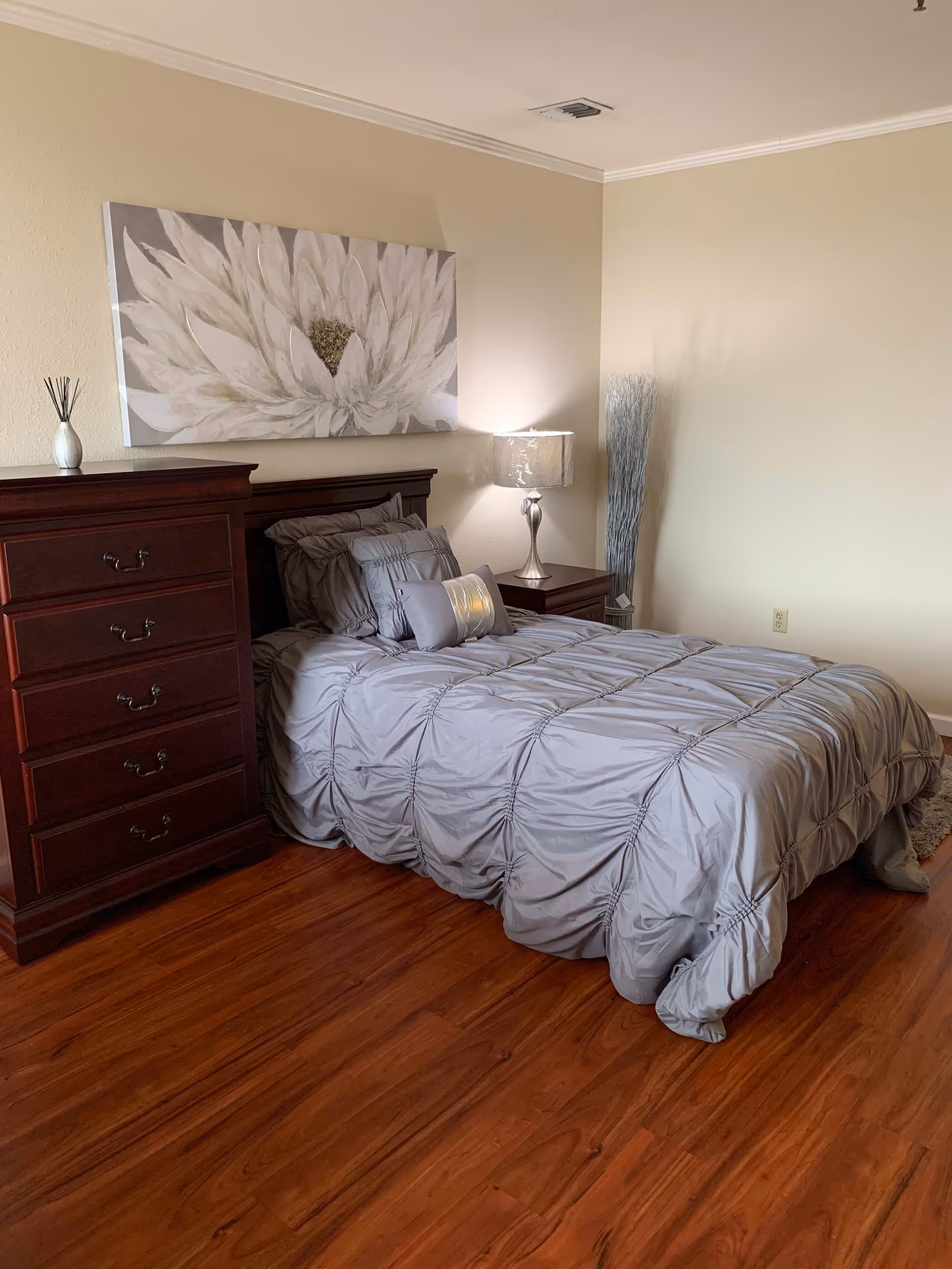 A bedroom with a wooden floor featuring a bed with a gray comforter and multiple pillows. Next to the bed is a wooden nightstand with a silver lamp and a tall decorative arrangement of sticks in a vase. On the left side of the bed is a wooden chest of drawers with a small decorative item on top. Above the bed hangs a large painting of a white flower.