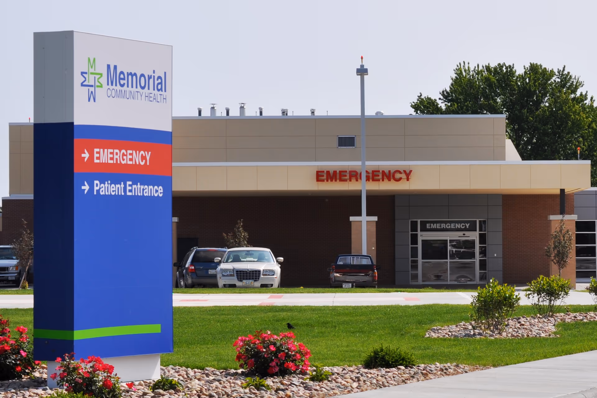 Exterior view of Memorial Community Health facility showing the emergency entrance with a large sign indicating directions for Emergency and Patient Entrance. There are cars parked near the building, green grass, and flower beds in the foreground.