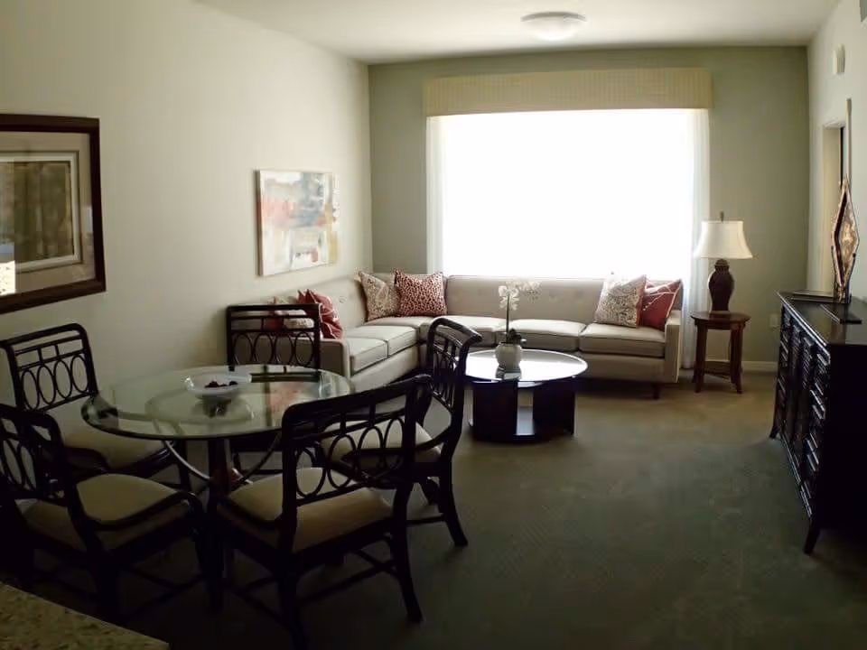 A cozy living room with a large window covered by a light-colored valance. The room features a beige L-shaped sofa adorned with patterned throw pillows, a round glass coffee table with a small plant, a wooden side table with a lamp, and a dark wooden cabinet. In the foreground, there is a glass dining table with four chairs that have cushioned seats and decorative backs. The walls are light-colored with framed artwork hanging on them.