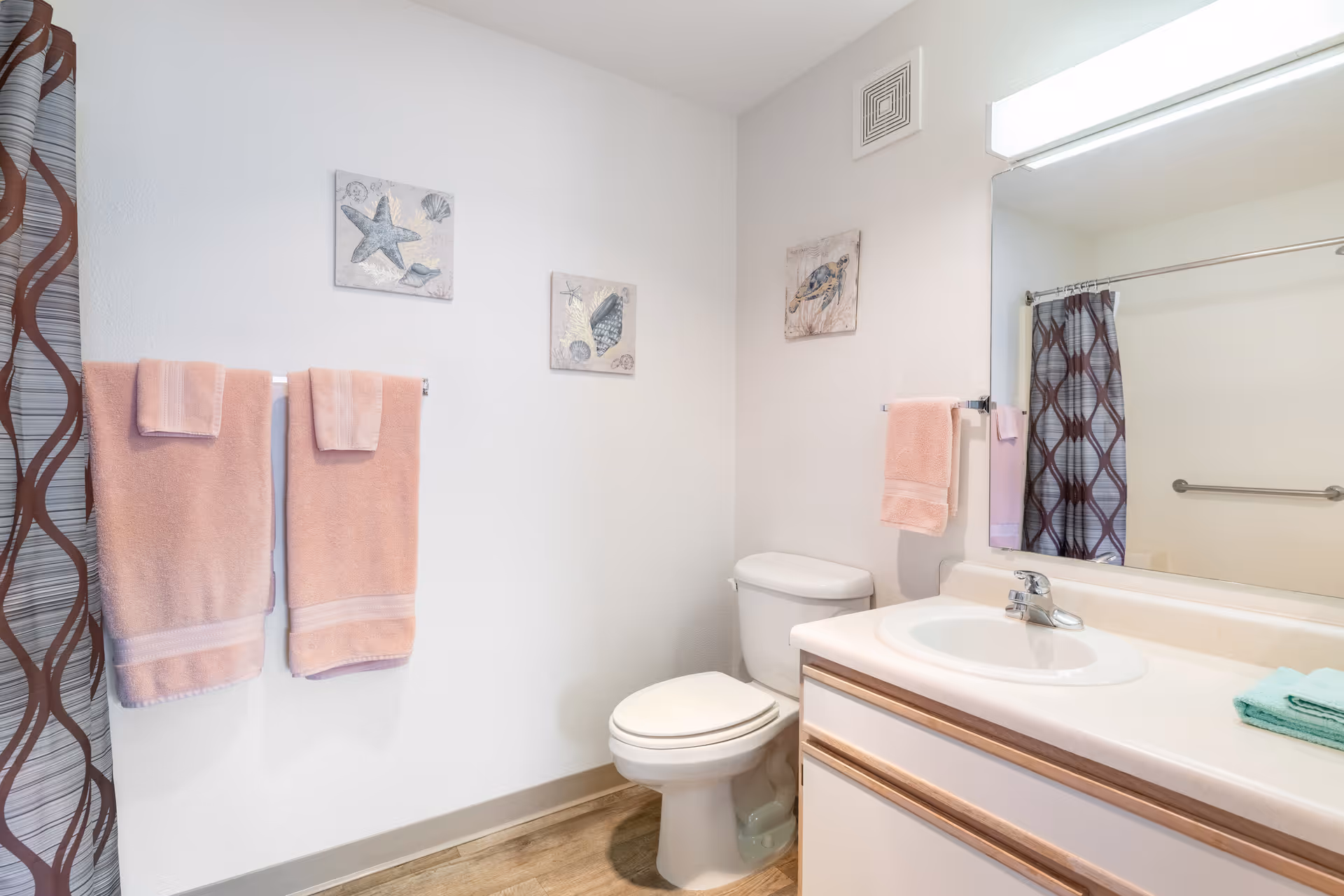 A clean and bright bathroom with a white toilet, a sink with a countertop, and a large mirror above the sink. There are two pink towels hanging on a towel rack on the wall and a smaller pink towel hanging next to the sink. The shower curtain has a brown and gray geometric pattern. The walls are decorated with three small framed pictures featuring sea creatures and shells.
