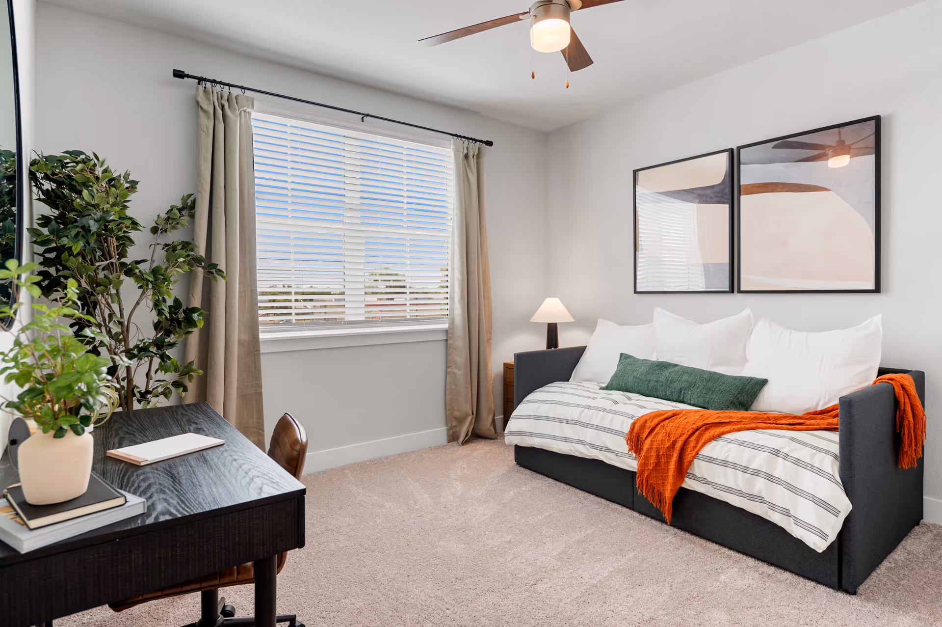 Bright furnished bedroom with a daybed, desk, window with blinds, and wall art.