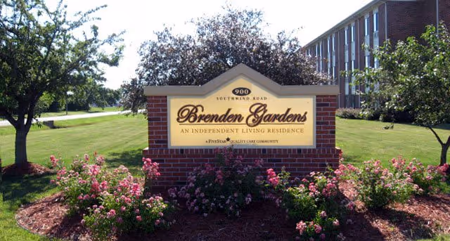 A brick sign for Brenden Gardens, an independent living residence, surrounded by landscaped flower beds with pink flowers and green bushes. There are trees and a multi-story brick building in the background under a clear sky.