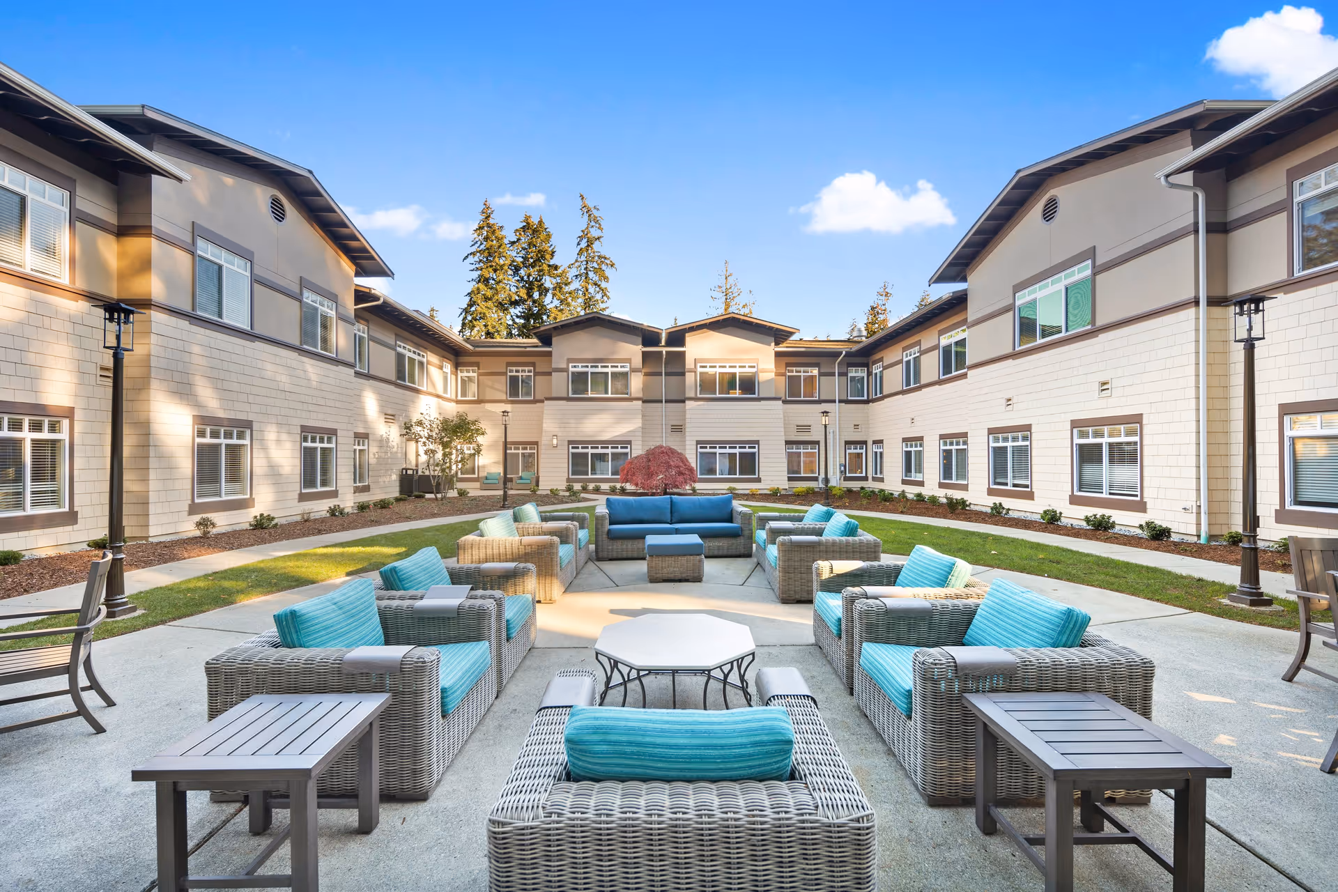 Outdoor courtyard area of a senior living facility with multiple wicker armchairs and sofas featuring turquoise cushions arranged around a central octagonal table. The courtyard is surrounded by a two-story building with many windows, under a clear blue sky with a few clouds.