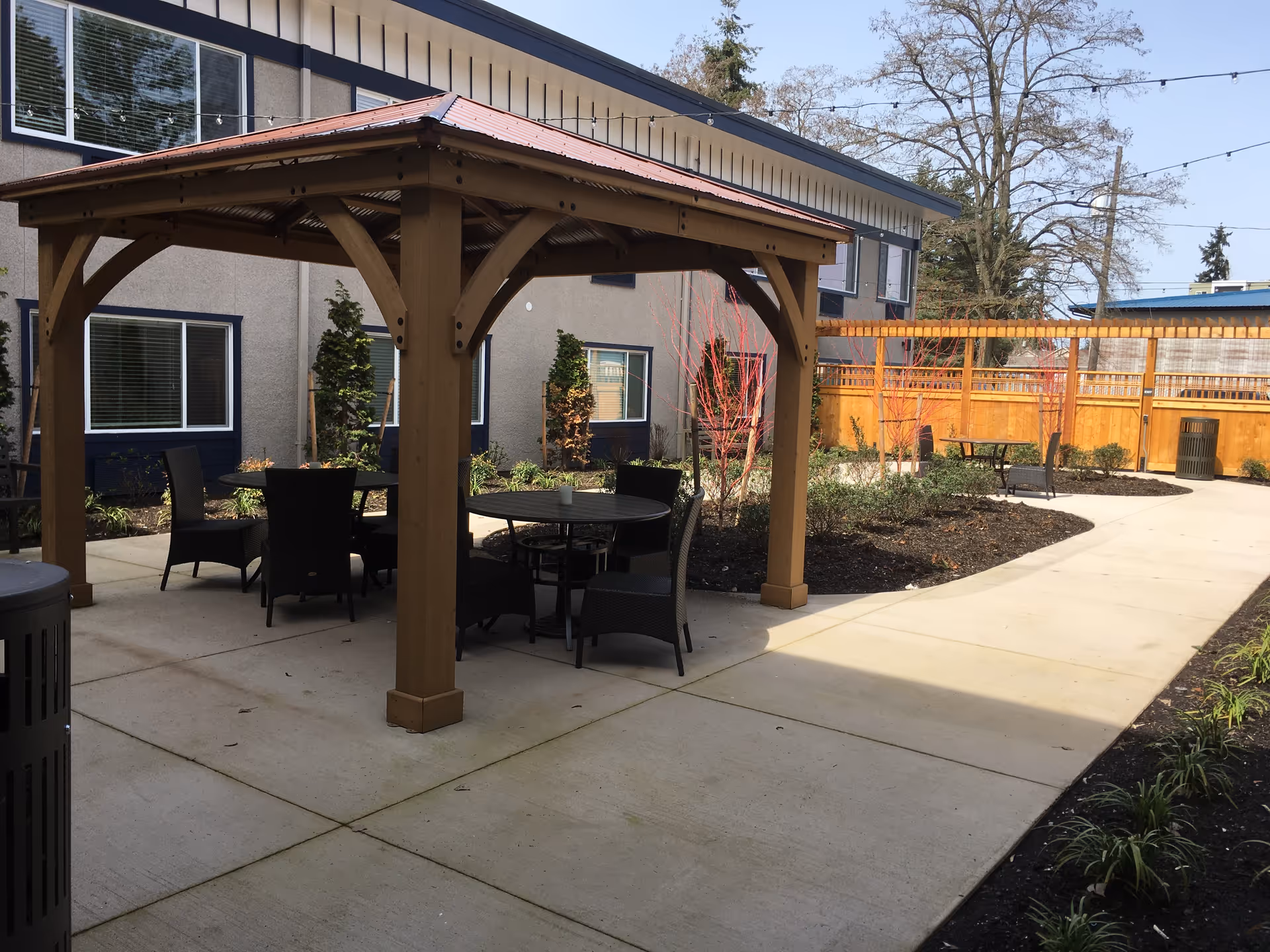 Outdoor patio area at a senior living facility with a wooden gazebo covering round tables and chairs. The area is surrounded by a concrete walkway, landscaped plants, and a wooden fence. The building exterior with windows is visible in the background under a clear sky.