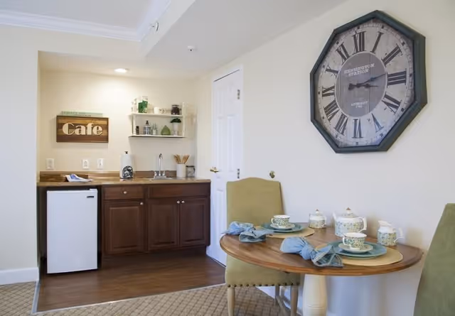 Small kitchenette area with dark wood cabinets, a mini refrigerator, a sink, and shelves holding decorative items. A wooden sign that says 'Cafe' is mounted on the wall. Adjacent to the kitchenette is a round wooden table set for two with green chairs, blue napkins, and floral-patterned tea cups and teapots. A large vintage-style wall clock hangs above the table on a light-colored wall.