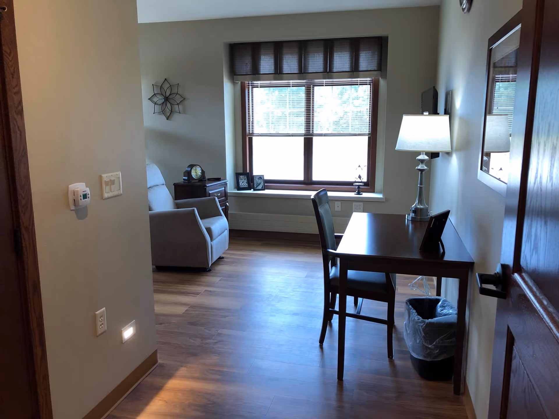 A cozy room with wooden flooring featuring a window with blinds, a wooden desk with a lamp and chair, a small trash bin, a comfortable armchair, a side table with a clock, and framed photos on the window sill.