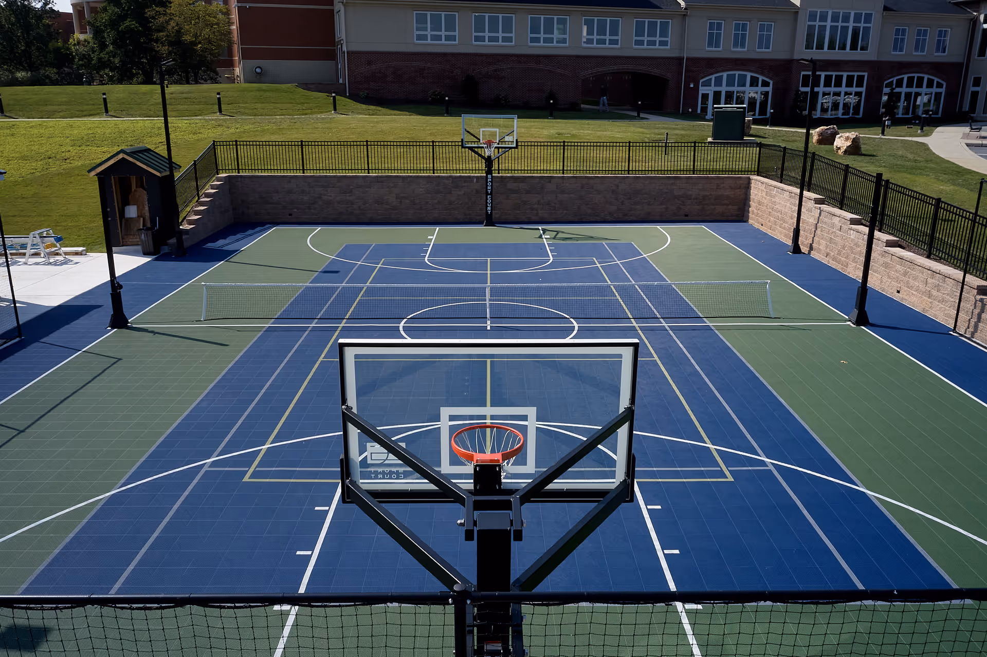 Outdoor multi-sport court with a basketball hoop, nets, and surrounding fence in front of a building.