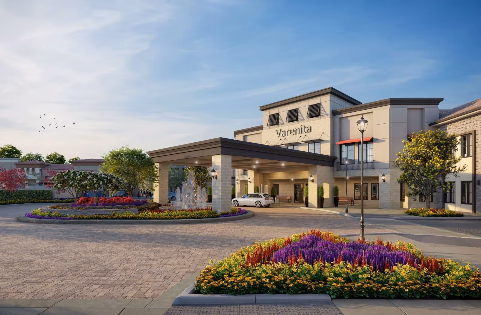 Exterior view of Varenita Simi Valley senior living facility showing the entrance with a covered drop-off area, a white car parked under the canopy, landscaped flower beds with colorful flowers, trees, and a clear blue sky with birds flying.
