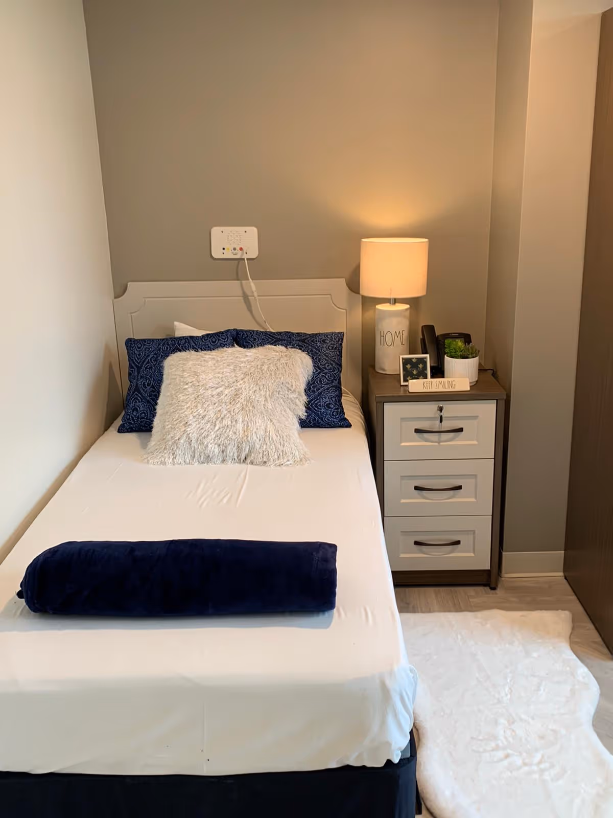 A neatly made single bed with white sheets, a dark blue rolled blanket at the foot, and three pillows including a fluffy white one and two dark blue patterned ones. Next to the bed is a nightstand with three drawers, a lamp with a beige shade, a small framed picture, a plant, and a decorative sign that says 'KEEP SMILING'. The wall behind the bed is painted gray, and there is a white rug on the floor beside the bed.