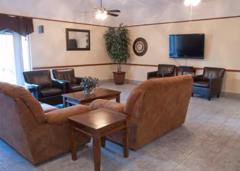 Cozy senior living lounge with sofas, chairs, plant, coffee table, and wall TV.