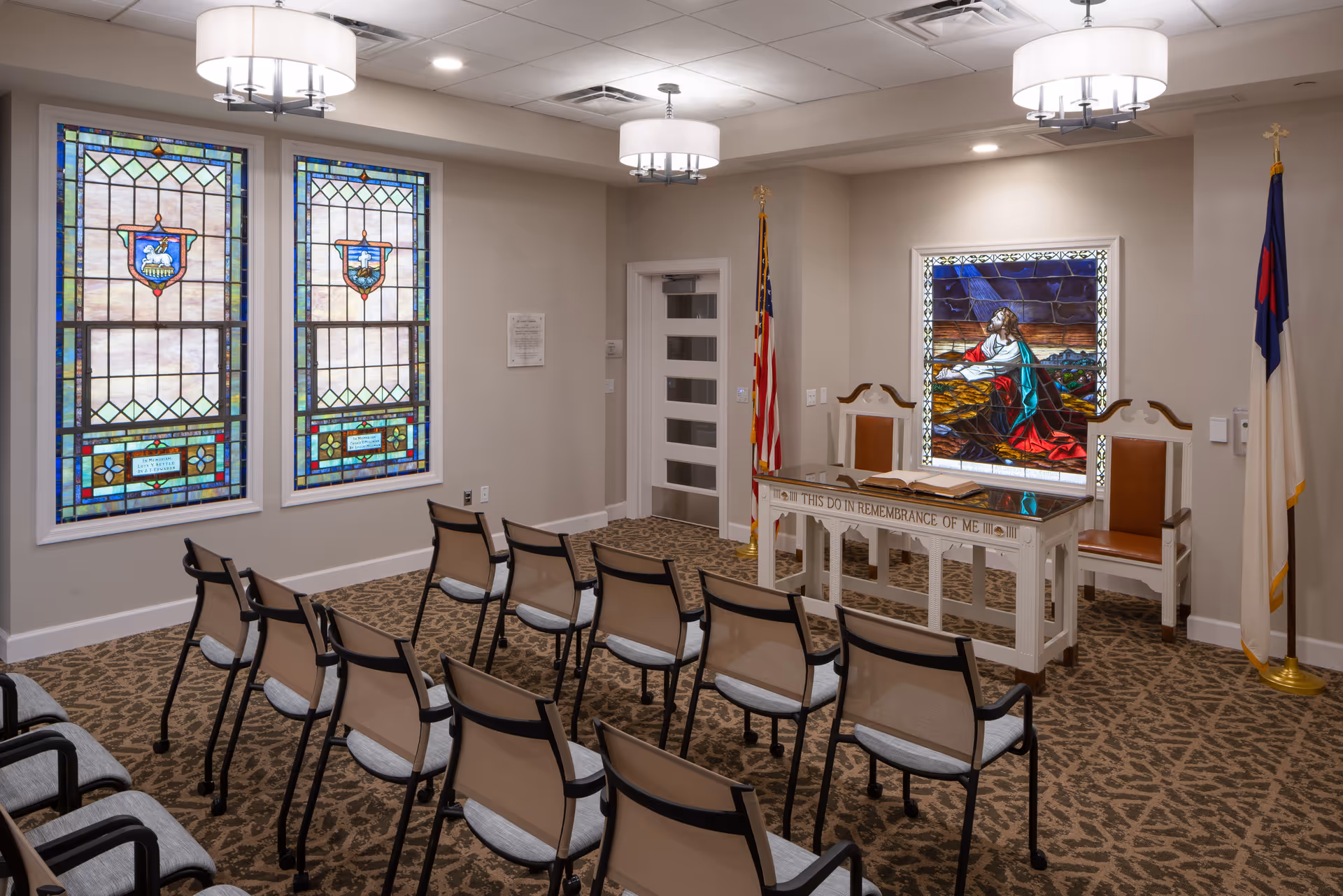 A small chapel room with rows of chairs facing a white altar table inscribed with 'THIS DO IN REMEMBRANCE OF ME'. Behind the altar is a stained glass window depicting a religious scene. Two additional stained glass windows are on the left wall. The room has beige walls, patterned carpet, and two flags placed near the altar.