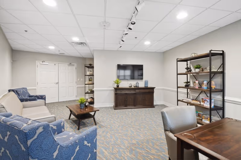 A well-lit living room area with a beige sofa, two blue patterned armchairs, a wooden coffee table with a small plant, a wooden cabinet with decorative items and a flat-screen TV mounted on the wall above it. There is a black metal bookshelf with various books and decor on the right side, and a wooden table with a gray chair in the foreground. The room has light gray walls, a patterned carpet, and a white ceiling with recessed lighting.