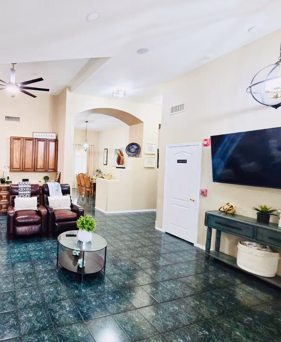 A bright and spacious living room area in Compassionate Manor Assisted Living featuring two brown leather recliners with cushions and throws, a round glass coffee table with a small green plant, a large flat-screen TV mounted on the wall, and a green console table with decorative items. The room has green tiled flooring, beige walls, a ceiling fan, and an archway leading to a dining area with a wooden dining table and chairs.