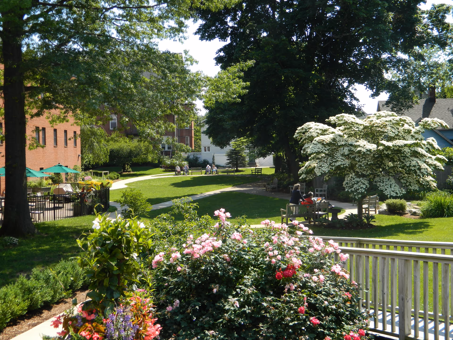 A sunny outdoor garden area at a senior living facility with green grass, blooming flowers, trees, and benches. Several people are sitting and walking along paved pathways. A brick building is visible on the left side with green umbrellas over tables.