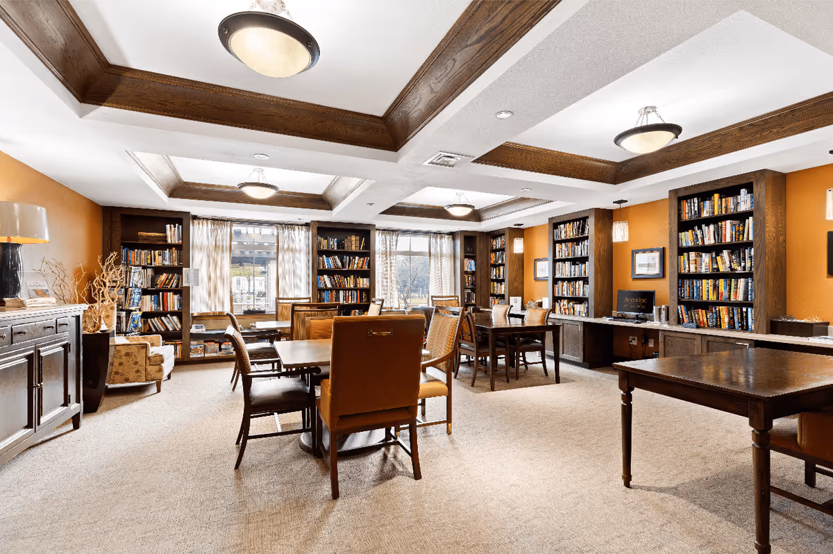 A bright and spacious library room with multiple wooden bookshelves filled with books along the walls. Several tables and chairs are arranged throughout the room for reading or studying. The ceiling features wooden beams and multiple light fixtures. Large windows with curtains allow natural light to enter the room.