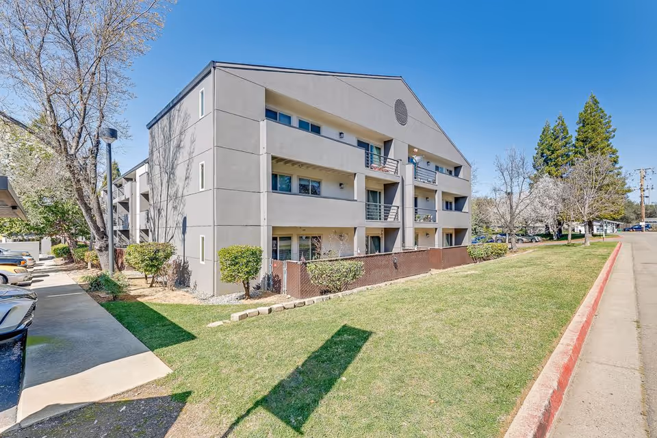 Exterior view of a three-story senior living facility building with balconies, surrounded by green grass, trees, and a clear blue sky. There is a sidewalk and parking area adjacent to the building.