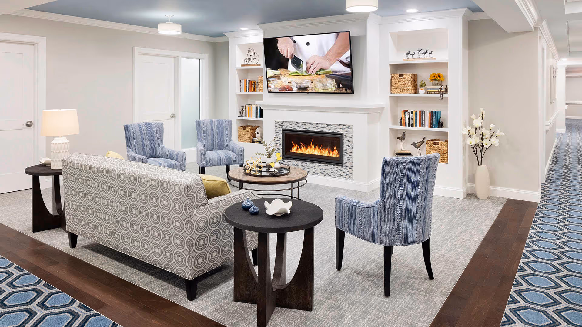 A cozy living room area in a senior living facility featuring a patterned sofa, three blue upholstered chairs, two dark wooden side tables, a round coffee table with decorative items, a modern fireplace with a fire burning, built-in shelves with books and baskets, and a flat-screen TV mounted above the fireplace showing a chef preparing food. The room has light-colored walls, a gray carpet, and a blue patterned hallway carpet visible to the right.