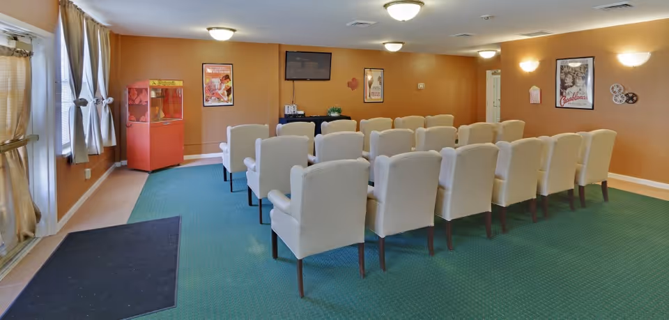 A small theater room with three rows of beige upholstered chairs facing a wall-mounted flat screen TV. The room has green carpet, tan walls decorated with vintage movie posters, a red popcorn machine in the corner, and windows with beige curtains on the left side.