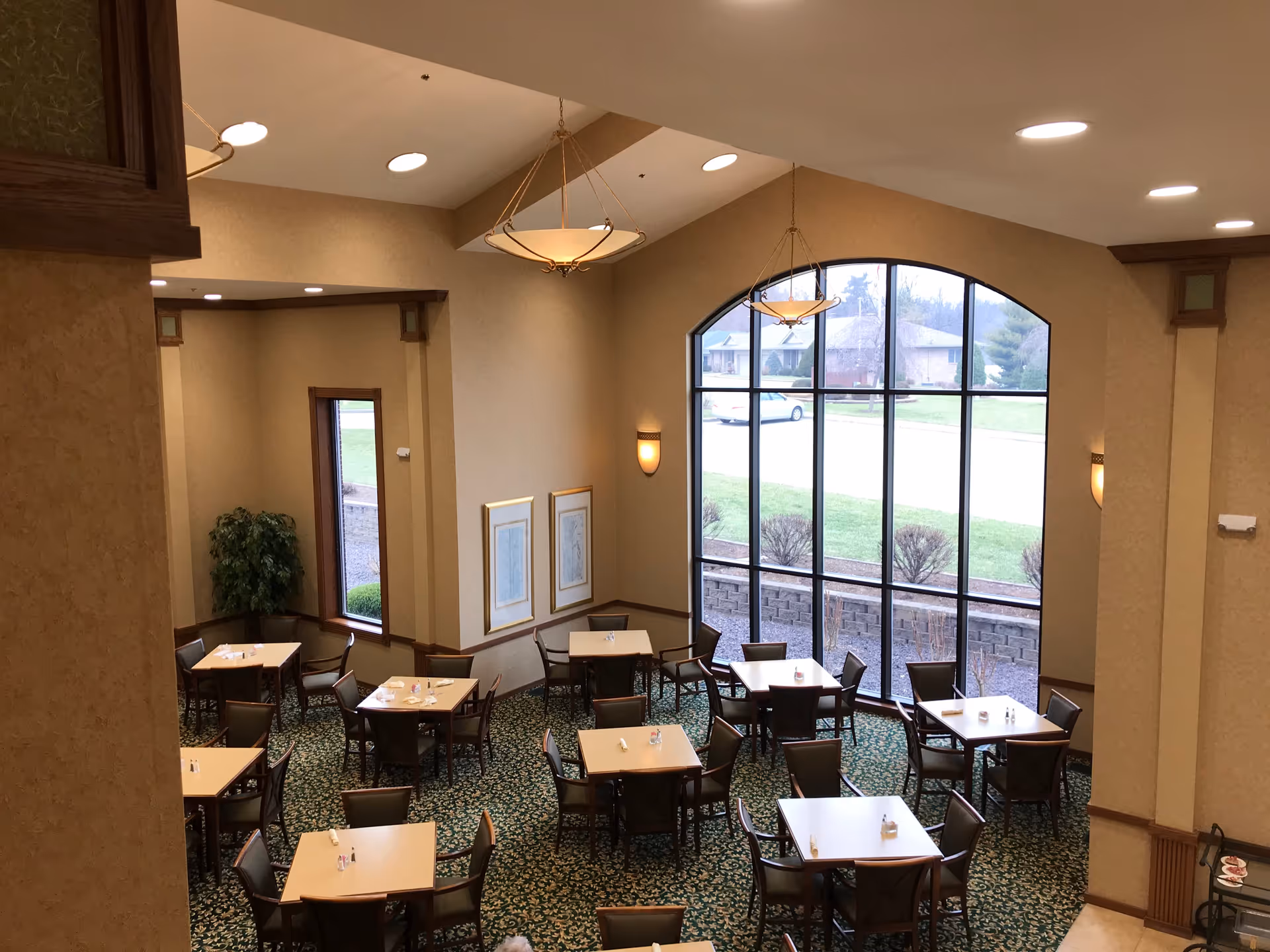 Interior view of a dining room with multiple square tables and chairs arranged neatly. The room has a high ceiling with hanging light fixtures and large arched windows letting in natural light. The carpet has a green patterned design and there are framed pictures on the walls.