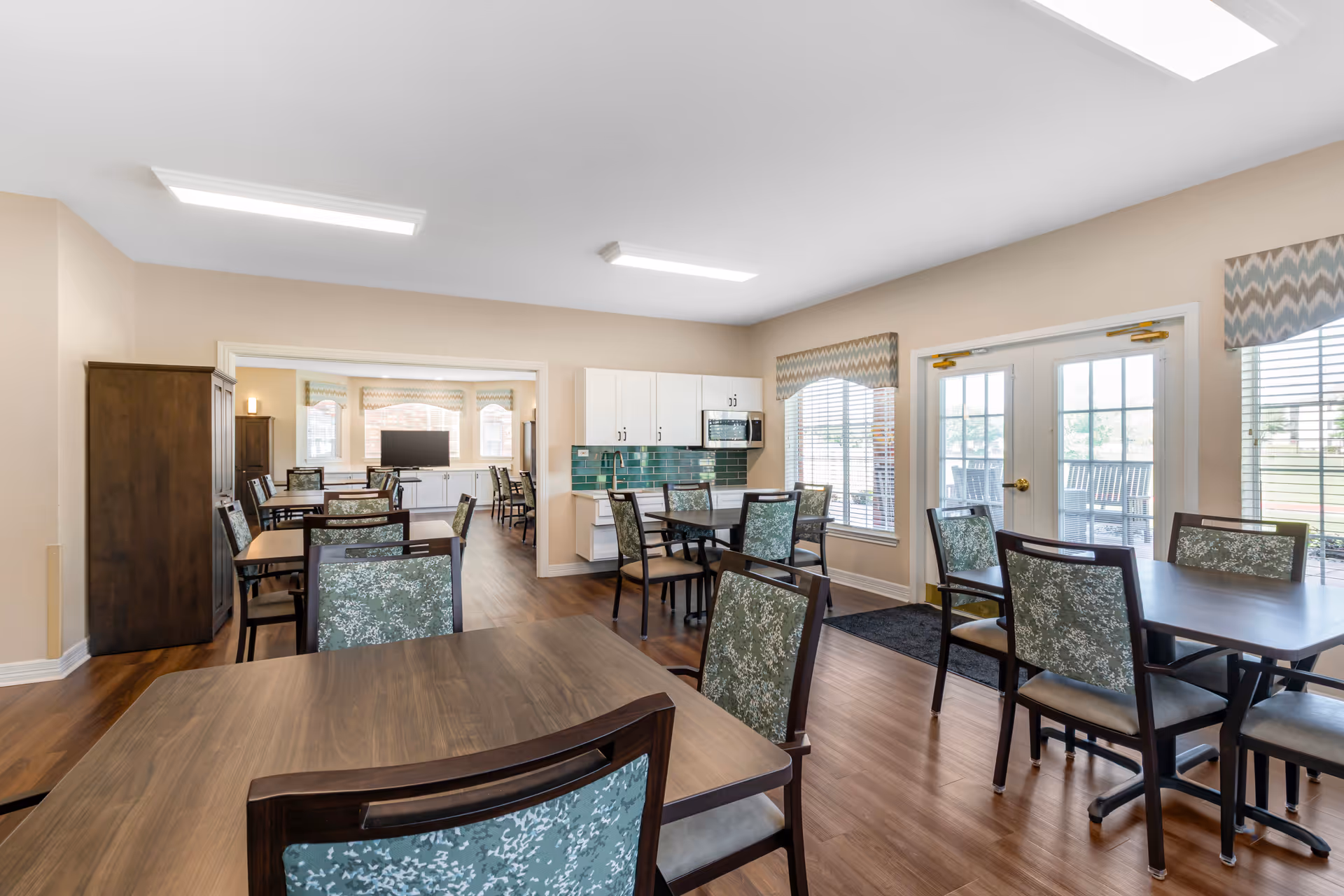 A bright dining room in a senior living facility with multiple wooden tables and chairs featuring green patterned upholstery. The room has large windows with chevron-patterned valances, a set of double glass doors leading outside, and a small kitchenette area with white cabinets and a microwave. The flooring is wood, and the walls are painted beige.