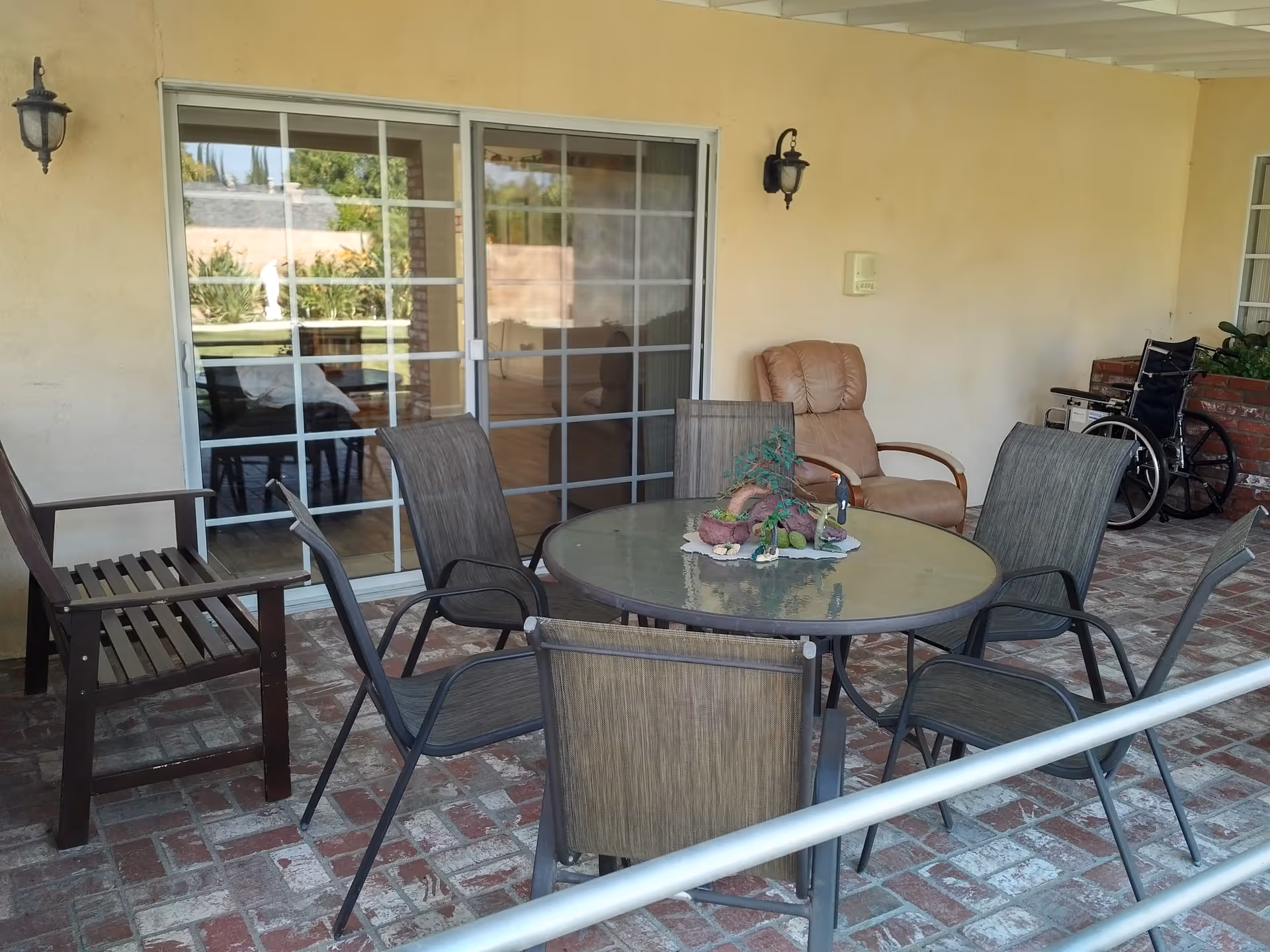 Covered outdoor patio area with a round glass table surrounded by six chairs, a wooden bench, a tan recliner chair, and a wheelchair near a brick planter. Sliding glass doors lead inside the building.