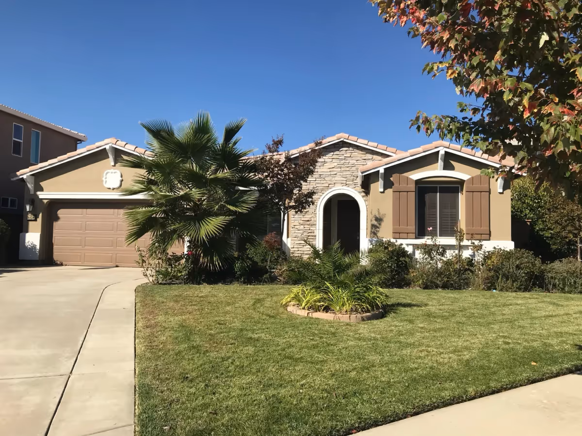 Single-story house with a stone and stucco exterior, a two-car garage, a palm tree, and a well-maintained lawn with shrubs and a tree with autumn-colored leaves under a clear blue sky.