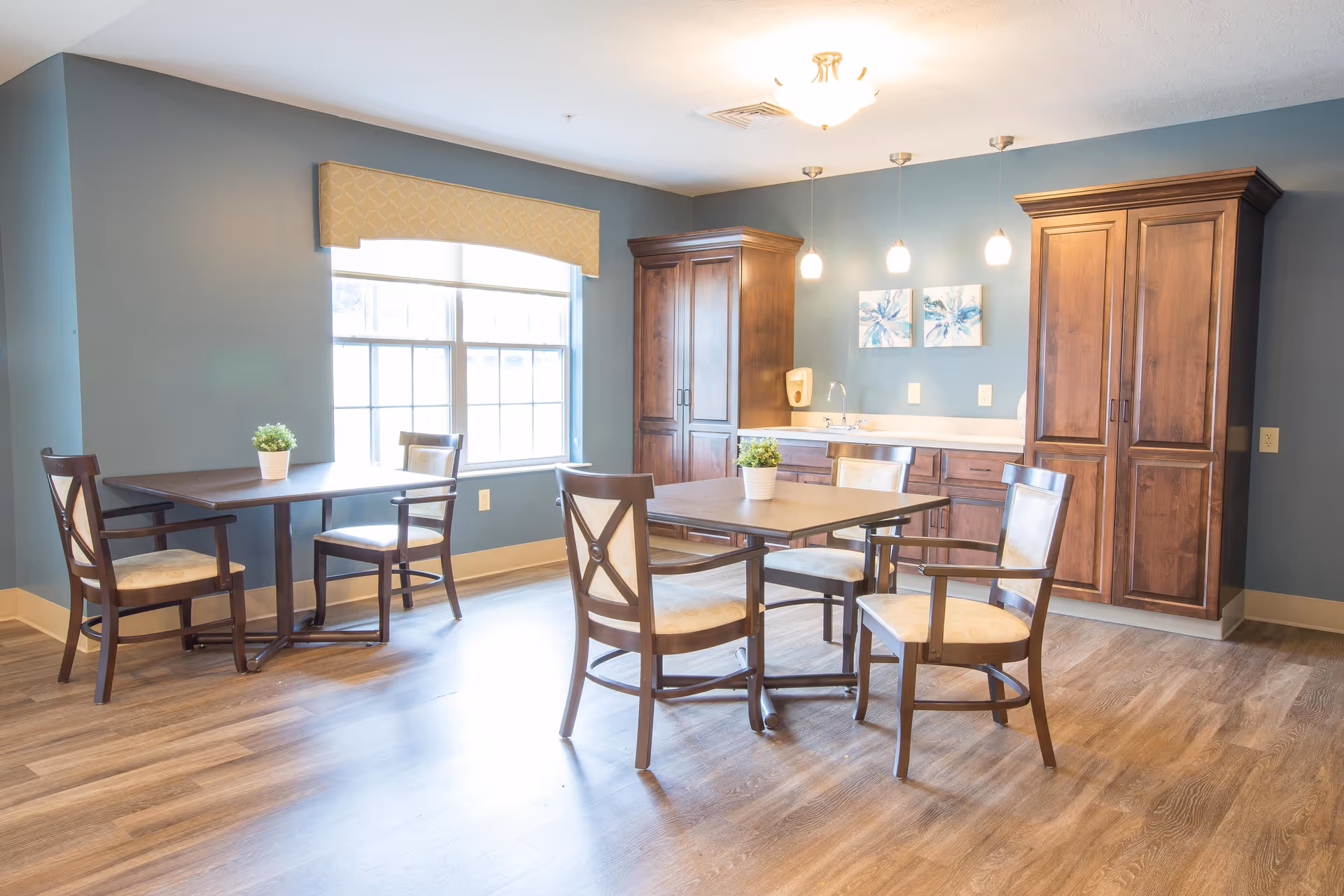 A bright dining area with two square tables, each surrounded by four wooden chairs with cushioned seats. The room has blue walls, a large window with a beige valance, wooden cabinets, a small sink, and two framed floral artworks on the wall. The floor is wood, and there are pendant lights hanging from the ceiling.