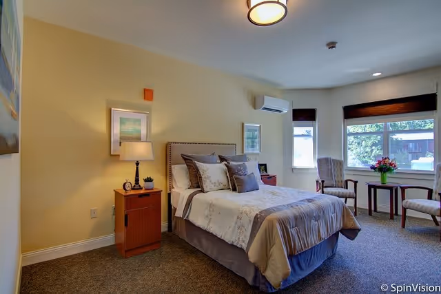 Furnished bedroom with a neatly made bed, nightstand with lamp, and seating by large windows.