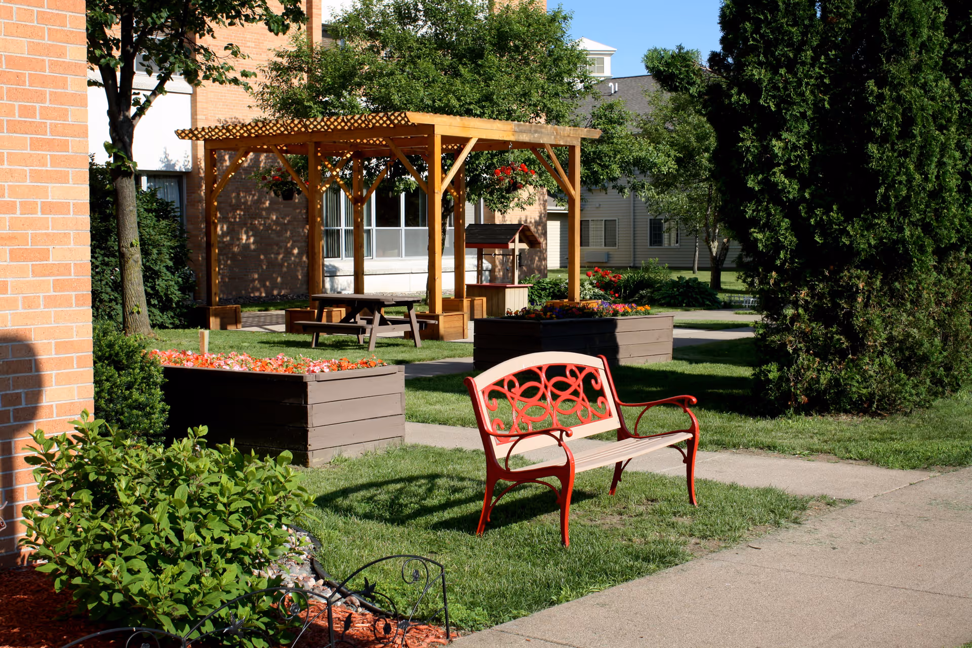 Outdoor garden area with a red and beige bench on grass, two large wooden planters filled with flowers, a wooden pergola with hanging flower pots, picnic tables underneath, and surrounding trees and shrubs near a brick building.