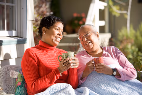 Two elderly women sitting outdoors on a bench, wrapped in a blanket, smiling and holding mugs while enjoying a sunny day.