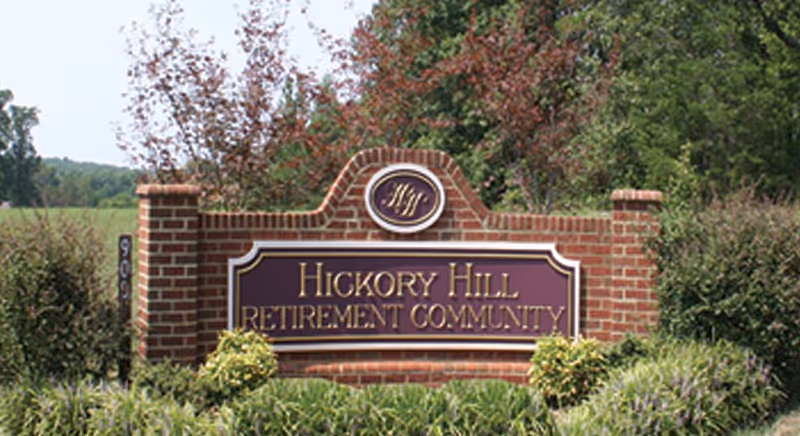 A brick sign with a purple and gold plaque that reads 'Hickory Hill Retirement Community' surrounded by green bushes and trees in the background.