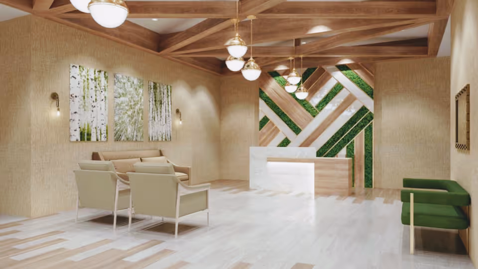 A spacious and modern interior lobby area with light wood flooring and beige textured walls. The ceiling features wooden beams with hanging round light fixtures. There is a white and wood reception desk with a decorative wall behind it featuring diagonal wood and green plant panels. On the left side, there are three beige armchairs arranged around a small sofa, and on the right side, there are two green cushioned chairs. Three nature-themed paintings hang on the left wall.